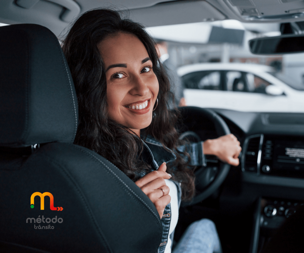 Mulher sentada no banco do motorista de um carro parado olhando para trás e sorrindo. Logo da Método Trânsito posicionada no canto inferior esquerdo.